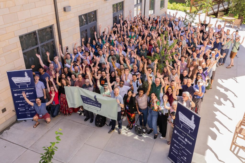 Group photo of State of the Map U.S. attendees outside the conference venue. Attendees are smiling and raising their arms expressing their excitement being a part of the event. The weather is sunny and warm.